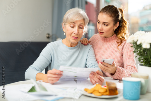 Thoughtful aged woman sitting at home table and reading documents while her adult daughter using cell phone to make copy of papers.