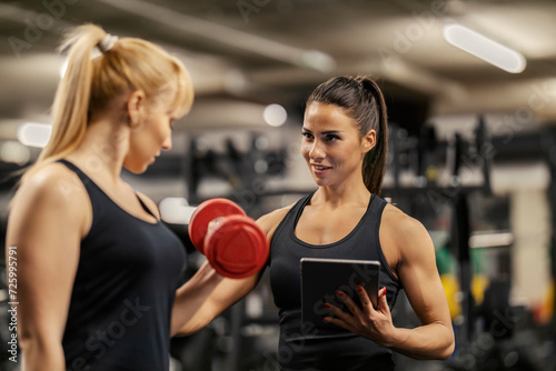 Portrait of a female coach with tablet training a sportswoman with dumbbells in a gym.