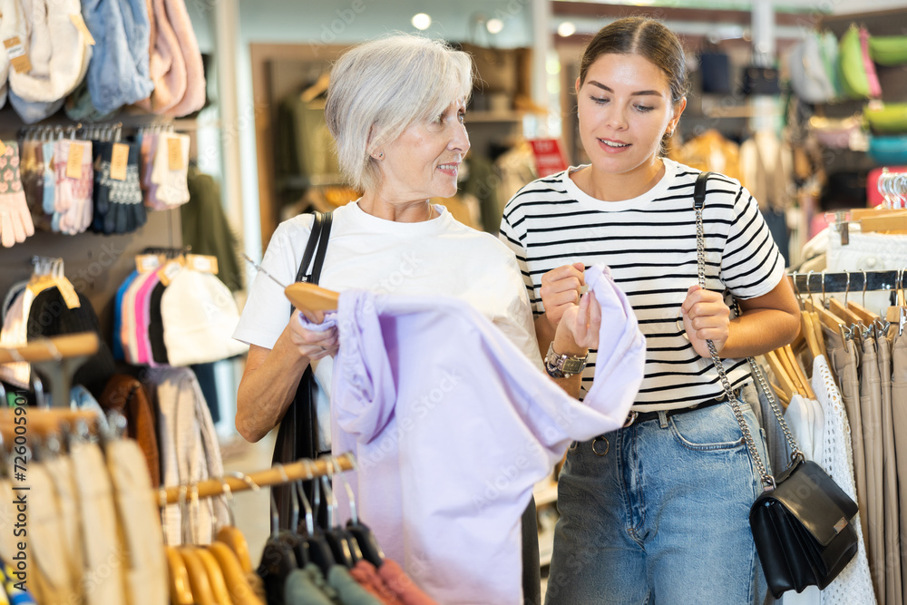 Obraz premium Young woman buyer in store has removed hanger with hoodie from window and lay heads together with old woman friend about purchasing. Girl imagine comfort and warmth that this coat will give