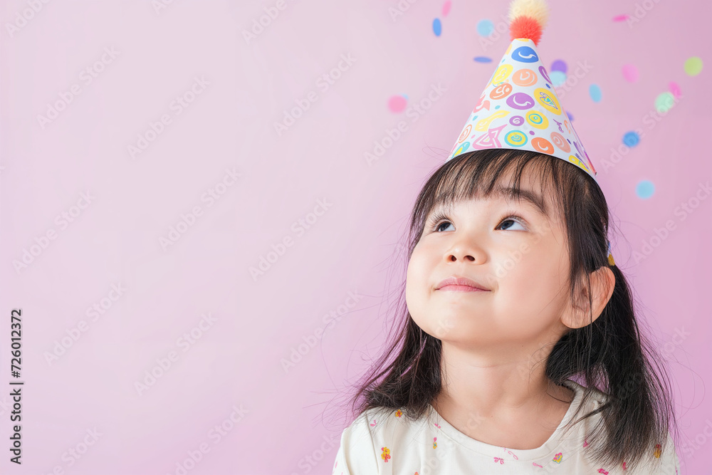 Smiling little asian girl looking up against a pastel background with a birthday party hat on her head Smiling little asian girl looking up against a pastel background with a birthday party hat on her head
