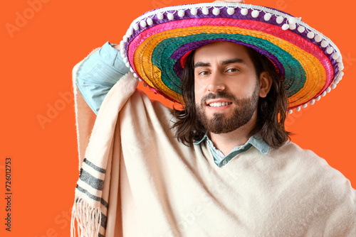 Handsome young Mexican man in sombrero and poncho on orange background