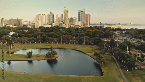 Designed park landscape at pond bank aerial. Green grass valley with flowers. Modern skyscrapers