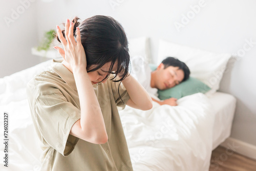 ベッドルームで耳を抑える女性と眠る男性（いびき・寝言・睡眠不足・ストレス・うるさい・睡眠障害） 