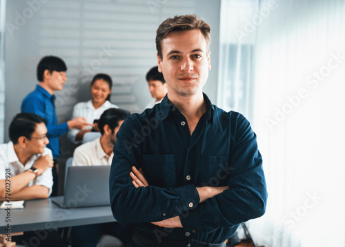 Confidence and happy smiling businessman portrait with background of his colleague and business team working in office. Office worker teamwork and positive workplace concept. Prudent