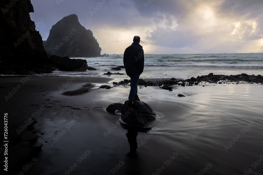 Man standing on sandy shoreline during a storm at sunset. Piha, Auckland, New Zealand.