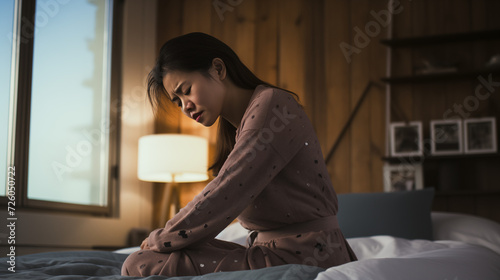 a young Asian woman sitting on a bed, holding her lower back and suffering from chronic back pain