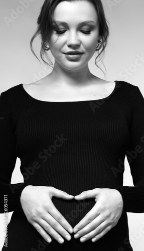 Black and white portrait of pregnant female in black dress with hands near pregnant belly with heart sign.