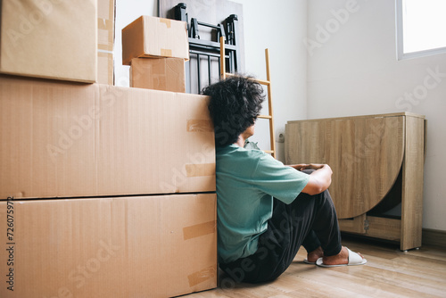 Young man sitting on floor leaning on pile of carboard boxes looking at window. Moving house concept photo
