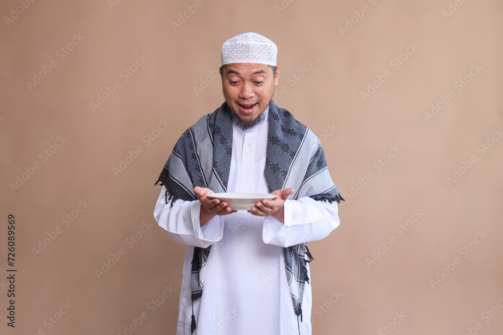 Excited Asian muslim man holding empty dinner plate on beige background Excited Asian muslim man holding empty dinner plate on beige background