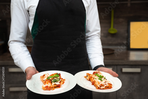 Waiter in Black Apron Serving Two Plates of Lasagna. A professional waiter in a black apron and white shirt holds two plates of lasagna garnished with fresh basil, showcasing the dish before service.