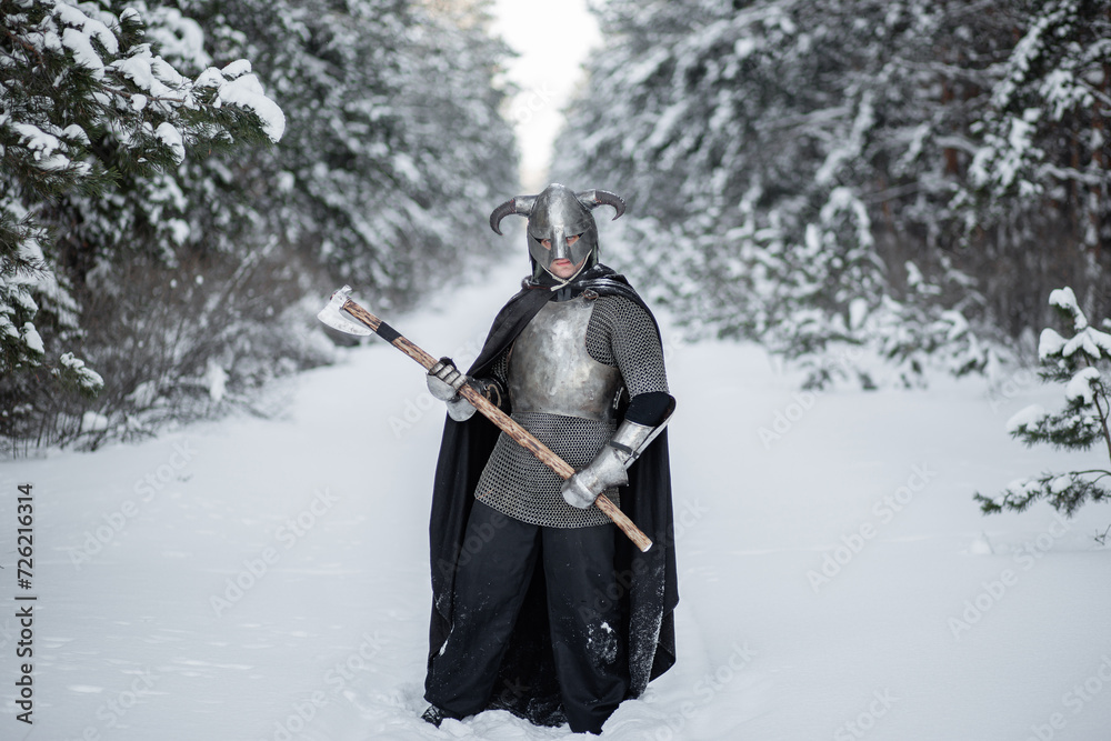 Naklejka premium Full-length portrait of a medieval fantasy warrior in a horned helmet, steel breastplate, chain mail with a two-handed ax in his hands, posing against the backdrop of a winter forest.