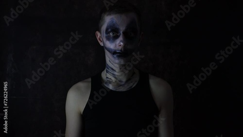 Young Caucasian man with a scary dark Halloween clown makeup costume standing in front of a black background photo