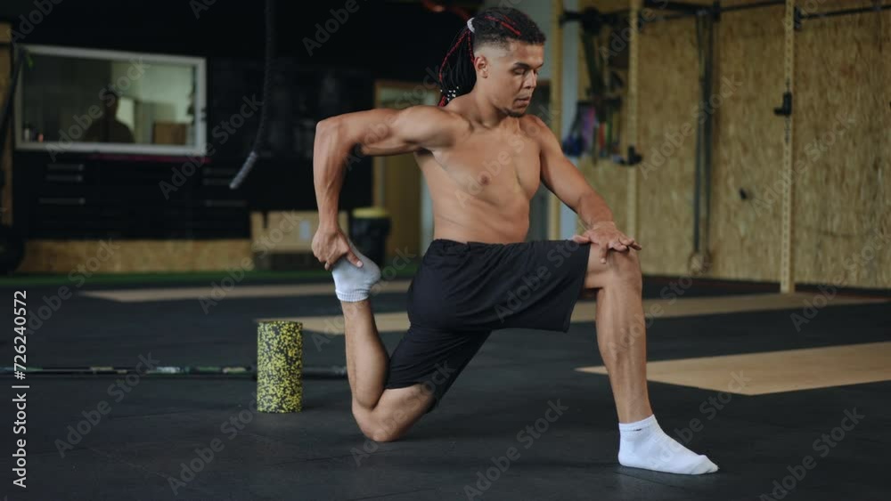 A mixed race man with dreadlocks on his head does a quadriceps stretch while standing on one knee in the gym A mixed race man with dreadlocks on his head does a quadriceps stretch while standing on one knee in the gym