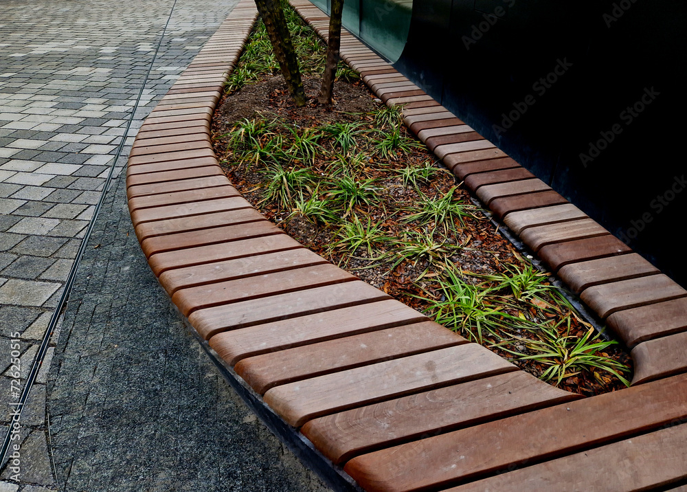drop shaped or ellipse bench on cobbled square. the paneling of  bench seating area is made of tropical teak wood with great resistance to wind and rain. multi stemed tree and ornamental grases
