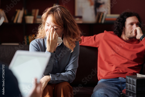 Selective focus of modern young woman wiping her tears with tissue during family therapy session photo