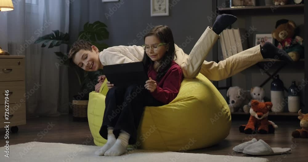 Little girl using digital tablet watching online video cartoon when preteen boy jumps on beanbag. Children using tablet for social media app together in the evening at home Stock ビデオ | Adobe Stock
