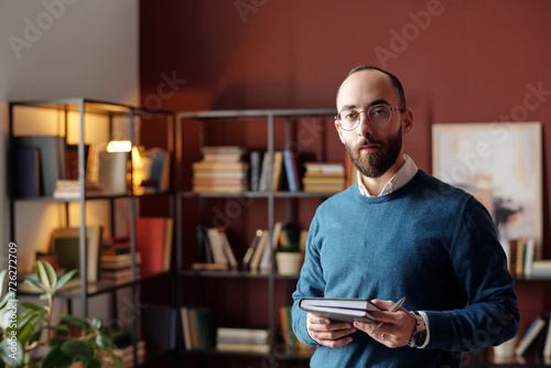 Medium portrait of young Middle Eastern psychotherapist with beard on face standing in office with notebook and pen in hands looking at camera photo