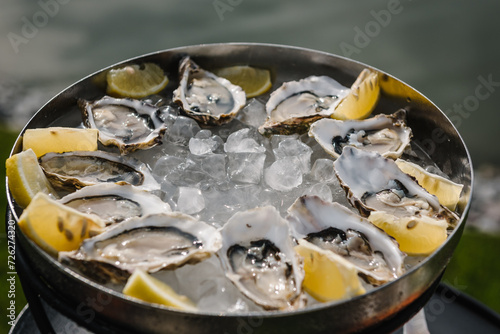 Oysters with lemon and ice closeup. Eating fresh live oysters at farm cafe in oyster farming village with view of water on sunny day. Food snack on table in pier or terrace restaurant.