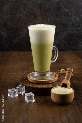 Ice matcha latte in a tall glass with wooden coaster and matcha green tea powder in wood table