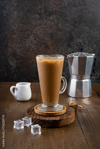 Ice coffee latte in a tall glass with wooden coaster and moka pot coffee maker