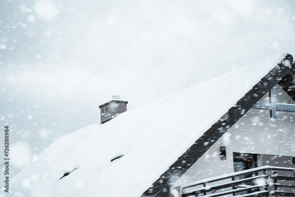 Smoke from a fireplace warms up a house in a winter wonderland as snow falls and covers the roof and the surrounding trees.