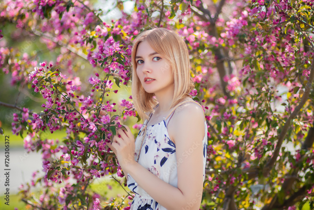 Portrait of an attractive young woman dressed in a dress posing against a background of blooming pink apple flowers. Portrait of an attractive young woman dressed in a dress posing against a background of blooming pink apple flowers.