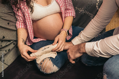 Unrecognizable pregnant woman at home wrapping baby bodysuit on floor while man is observing. Concept of first-time parents.