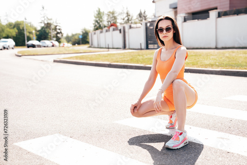 Young beautiful confident female in trendy summer cycling shorts and tank top clothes. Carefree woman posing in street. Positive model having fun. Cheerful and happy. Sits on asphalt
