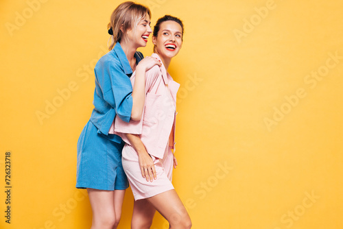 Two young beautiful smiling brunette hipster female in trendy summer clothes. Sexy carefree women posing near yellow wall in studio. Positive models having fun. Cheerful and happy