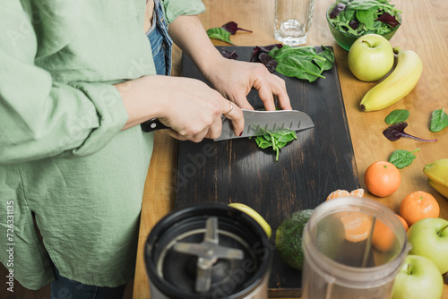 Woman cutting green spinach leaves, preparing healthy vegan smoothie with spinach, banana, apple and avocado in a blender on a table