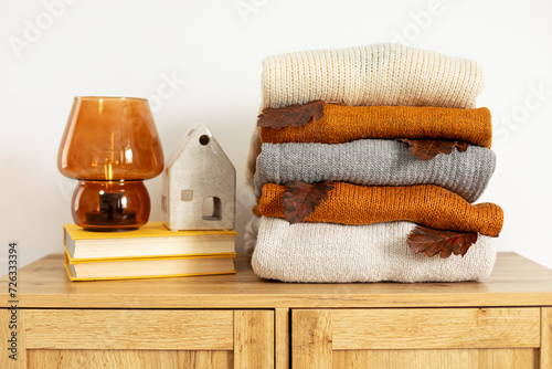 Stack of cozy knitted warm sweater with fall leaf, books, decorative broun lamp and ceramic house. Cozy hygge concept. Home autumn composition in the interior of the room. Simple minimal home cozy. photo