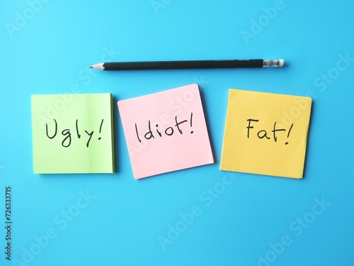 Paper on blue background with pencil writing bully words UGLY FAT IDIOT, concept of someone who hurts or frightens victims by degrade or demean in some way to feel powerful or stronger photo