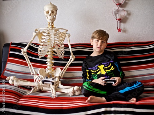 Young boy dressed as a coloured Halloween skeleton holds a joystick sitting on the sofa with skeleton playing video games photo