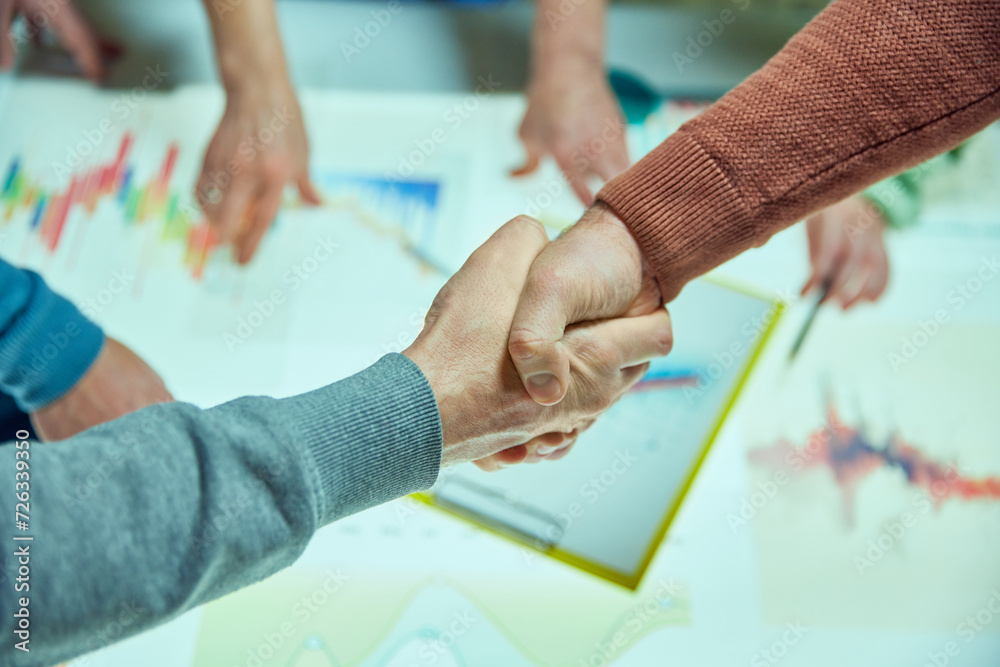 Two people shaking hands over a table with business graphs, symbolizing agreement and partnership. Start of new project and cooperation. Concept of business, teamwork, modern office