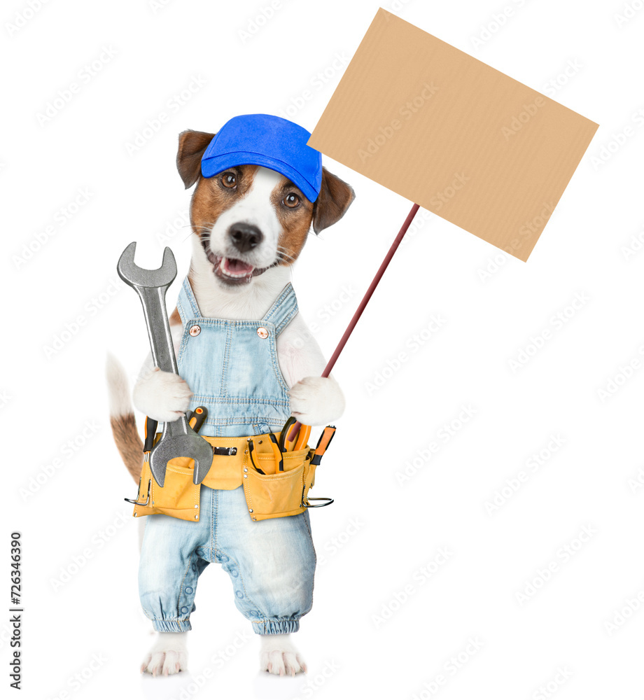 Funny Jack russell terrier puppy wearing red cap and denim overalls with tool belt holds wrench and shows empty placard. isolated on white background