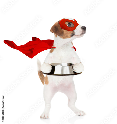 Funny jack russell terrier puppy wearing superhero costume holds empty bowl and looks away on empty space. Isolated on white background