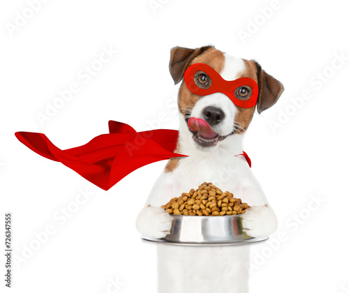 Licking Jack russell terrier puppy wearing superhero costume holds bowl of dry dogs food. Isolated on white background