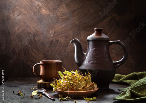 Still life with dried linden blossoms in a plate and linden tea in a teapot on a dark background