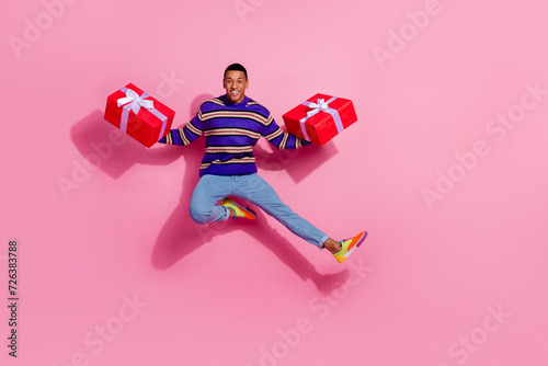 Full length photo of cool excited man wear striped sweater jumping high holding two present boxes empty space isolated pink color background