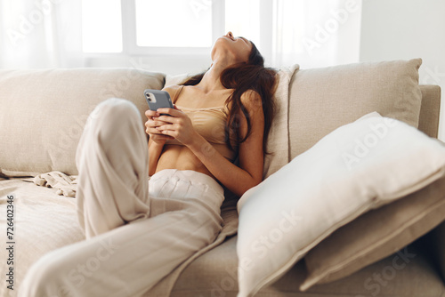 Cozy Home Relaxation: Beautiful Woman Holding Smartphone, Smiling and Chatting Online, Sitting on Sofa in Living Room.