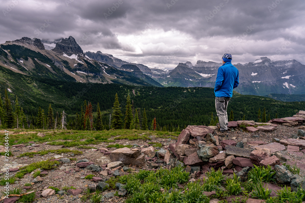 Man standing watching a scene of a mountain range with snow capped peaks under heavy clouds, Granite Park, Glacier National Park, Montana