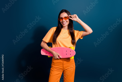 Photo of pleasant woman dressed orange apparel touching sunglass holding her new cool skateboard isolated on dark blue color background