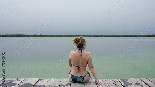 woman in bikini looking at green lagoon sitting on wooden pier