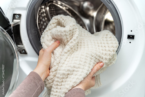 Woman putting white wool sweater into the drum of a washing machine in laundry room. 