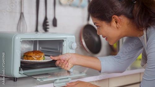 Indian woman cooking burger by placing it on microwave oven at kitchen - concept of healthy eating, domestic lifestyle and technology photo