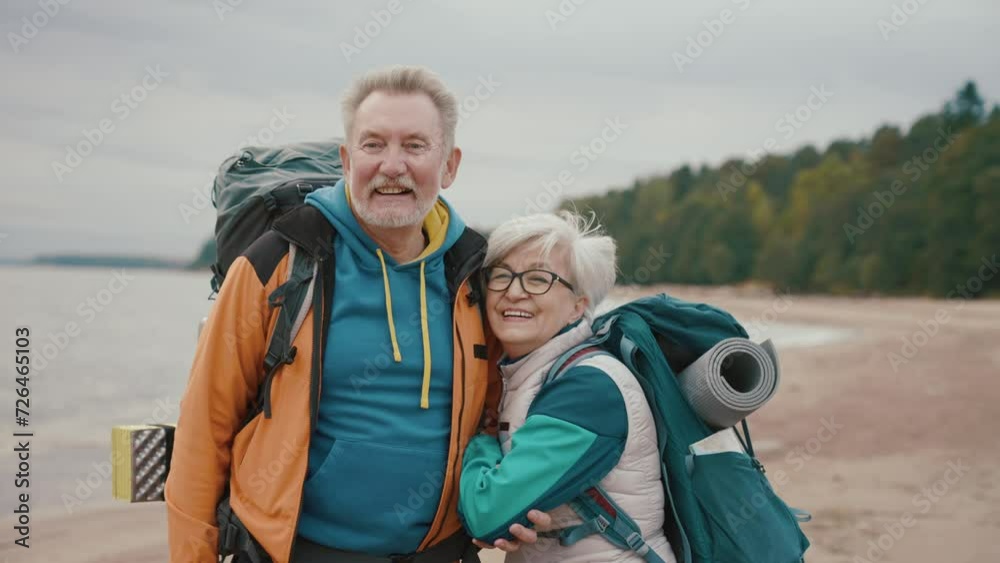 Portrait senior couple travelers hikers carrying backpacks with touristic equipment standing on riverbank looking at camera. Man woman pensioners hiking together. Tourism, freedom, hiking concept. Portrait senior couple travelers hikers carrying backpacks with touristic equipment standing on riverbank looking at camera. Man woman pensioners hiking together. Tourism, freedom, hiking concept.
