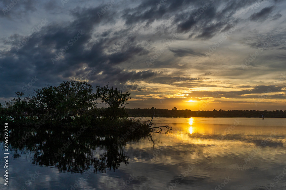 The setting sun is reflected in the Tomoka River, silhouetting a mangrove island. The trees and clouds are reflected in the water in this dramatic landscape. Photographed in Tomoka State Park, FL.