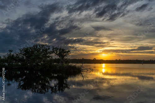 The setting sun is reflected in the Tomoka River, silhouetting a mangrove island. The trees and clouds are reflected in the water in this dramatic landscape. Photographed in Tomoka State Park, FL.