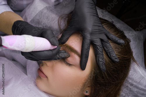 Close-up of the hands of the master in black gloves who holds the model's eyebrow and applies permanent makeup. PMU Procedure, Permanent Eyebrow Makeup.