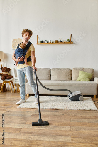 curly father with infant baby boy in carrier vacuuming hardwood floor living room, housework photo